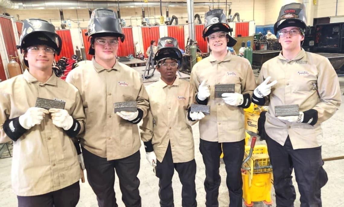 Associates in welding gear holding completed practice welds.