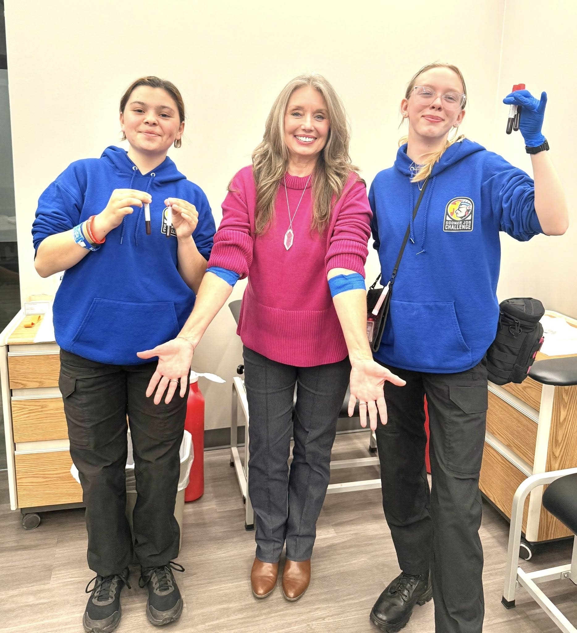 Two Phlebotomy Associates take a group picture with the SJC Director. Students are holding vials of blood, director has arms bandaged after volunteering as a practice subject for blood draws.