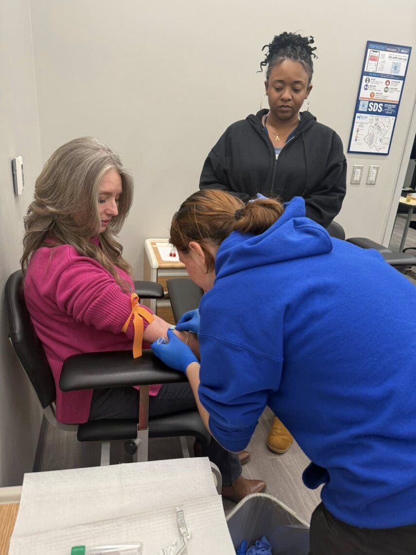 Associate inserts blood drawing needle into Ms. Master's right arm while instructor supervises. 
