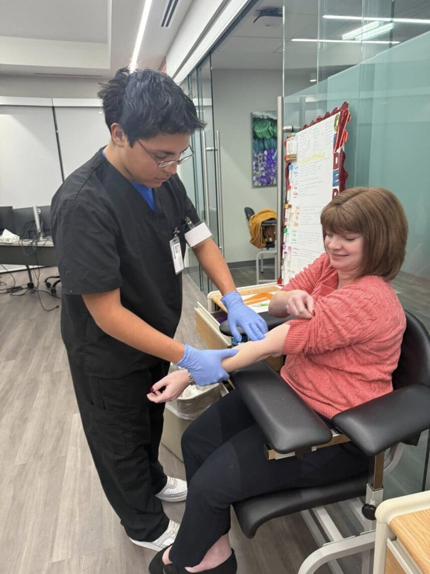 Associate prepares Ms. Meders for her blood draw. Woman is seated in chair while Associate stands over her in scrubs and gloves, holding and pressing on her arm.