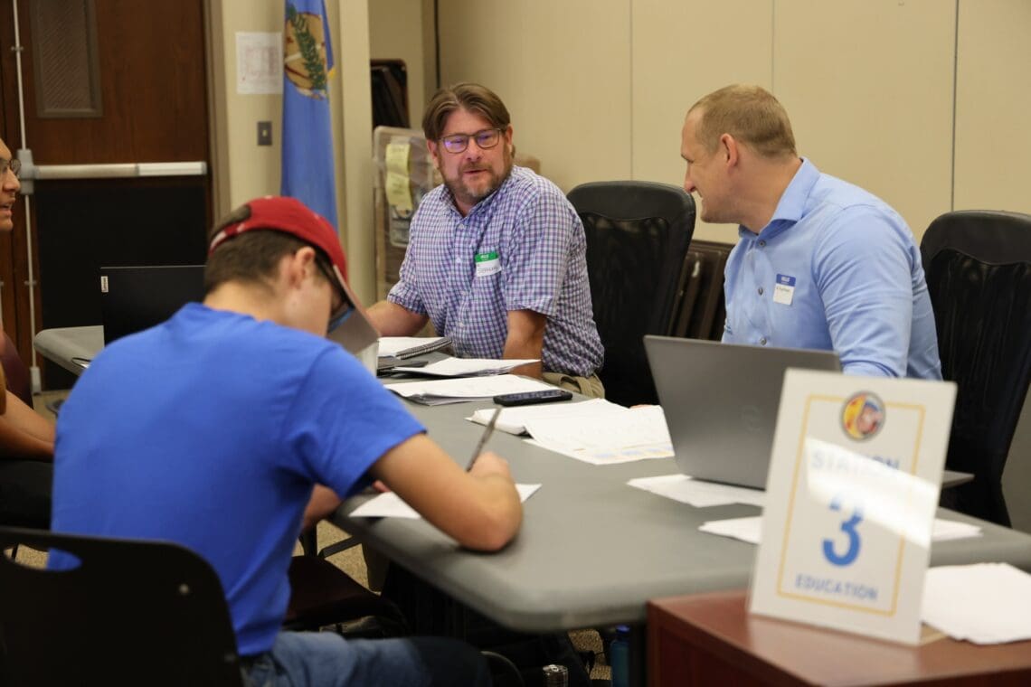 Two staff members talk to each other. They are sitting at a table while an Associate fills out paperwork on the other side of the table. There is a sign that says "Station 3: Education"