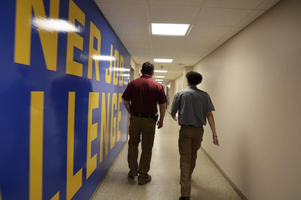 Two staff members walk together down a hallway, backs toward the camera. There is a mural on the wall to their right with the SJC logo on it. 