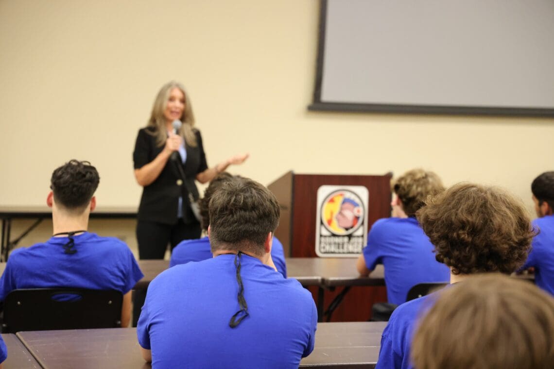 Program Director stands in front of group of seated associates with microphone, welcoming them to the program. - taken from behind Associates.