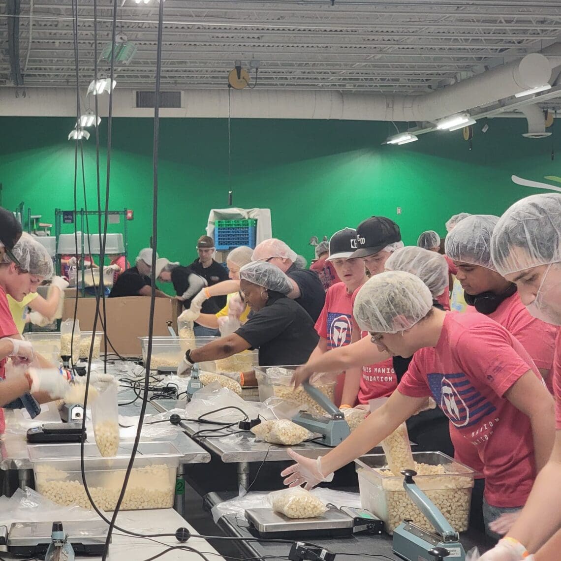 A group of SJC Associates and Staff measure and package food at the Regional Food Bank. They are all wearing Travis Manion Foundation tshirts and hairnets. 