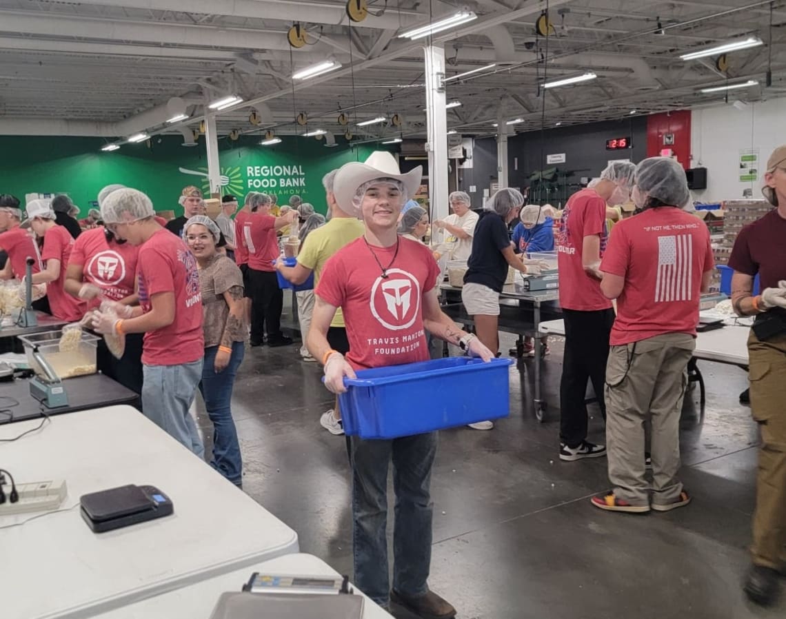 Associates are volunteering at Regional Food bank, completing various tasks around the room. In the center of an image, one Associate, who is wearing a hairnet and a cowboy hat, smiles while holding a blue tub.