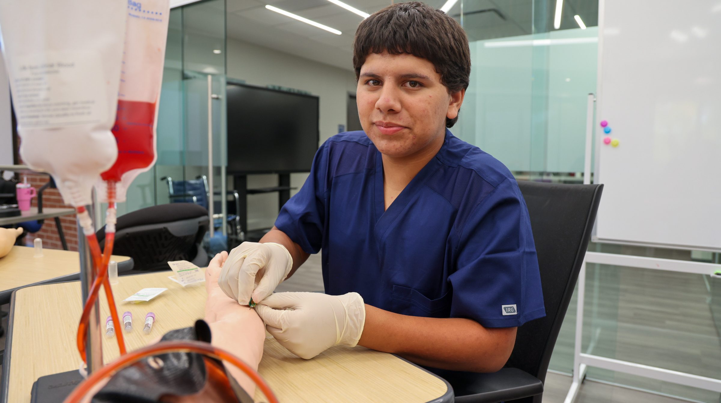Male phlebotomy student putting a butterfly needle in the arm of a dummy arm and smiling at the camera.