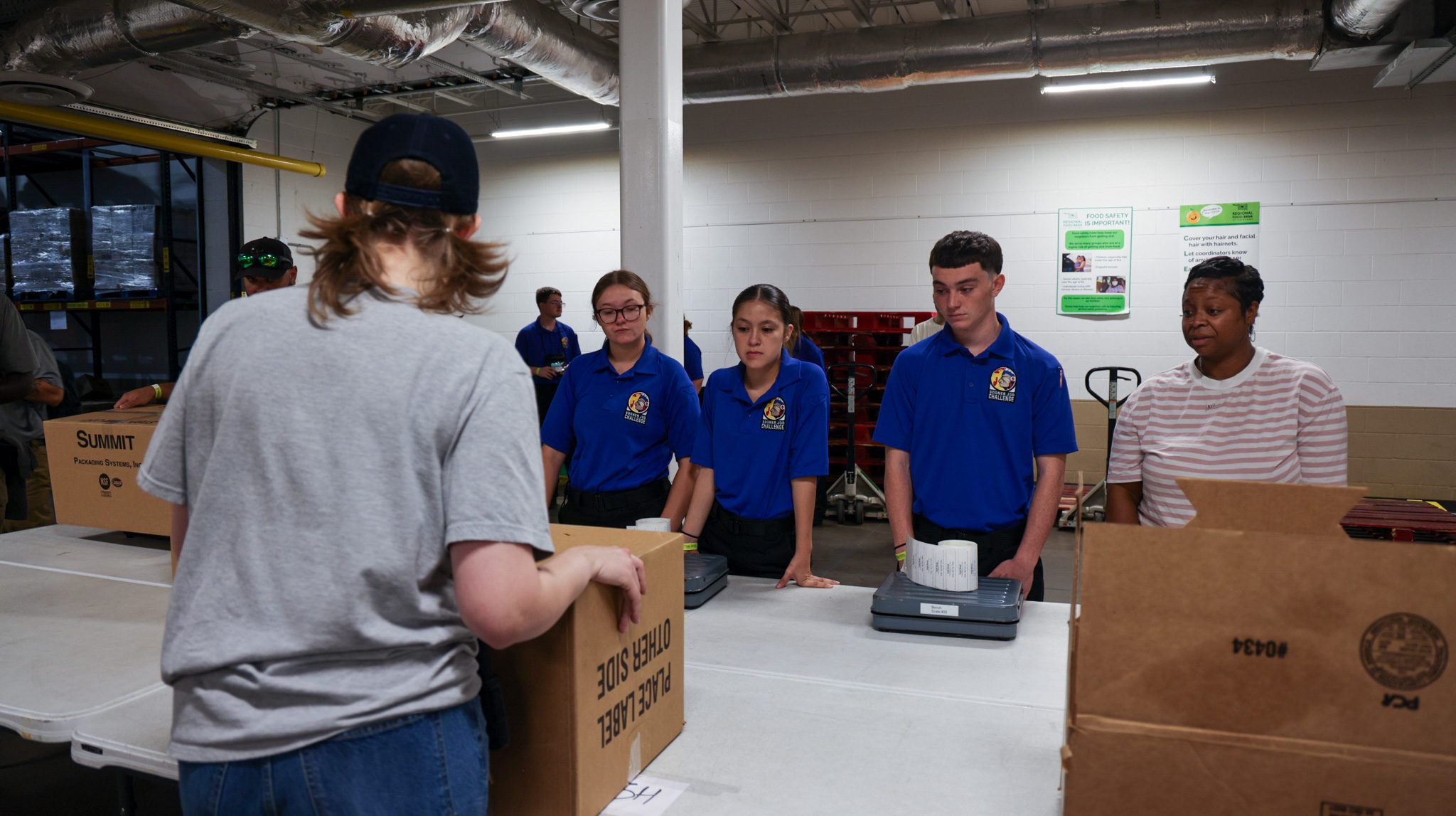 A group of students and one woman getting directions about packing the boxes in front of them.