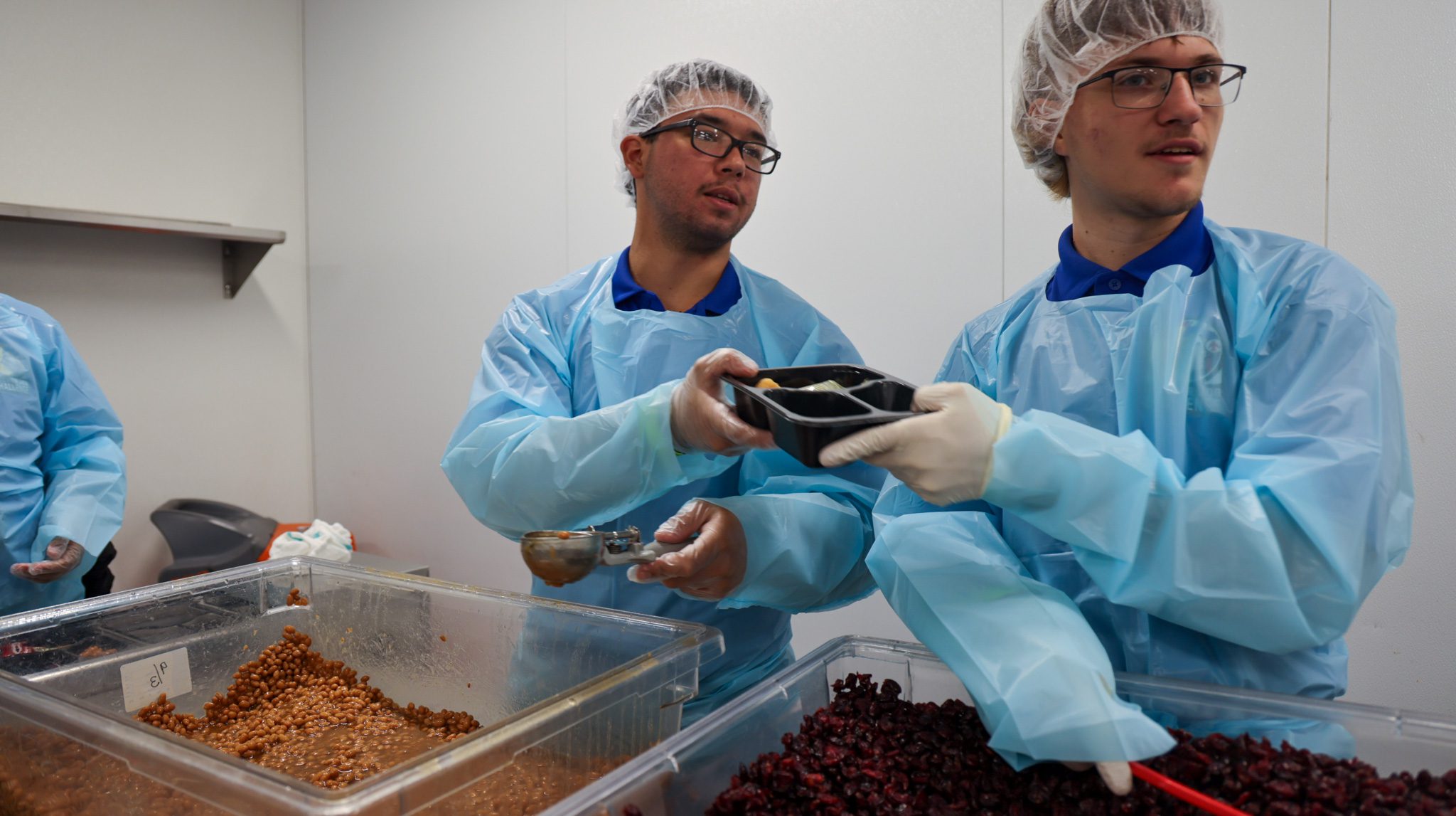 Two male students in protective clothing putting food into single-serving containers.