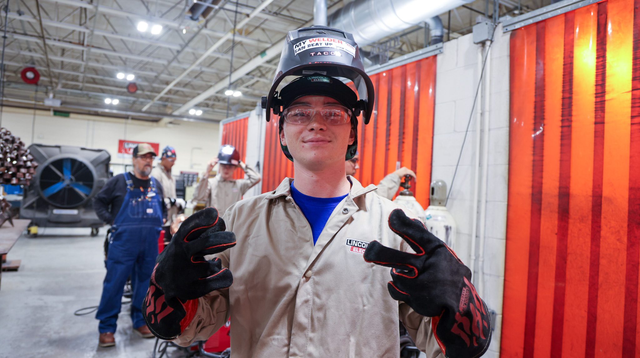 Male student welder with his helmet up holding up the peace sign and smiling.