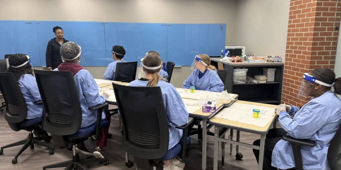 Class of Phlebotomy students sit in a group around a table while instructor speaks. Students are wearing. PPE.