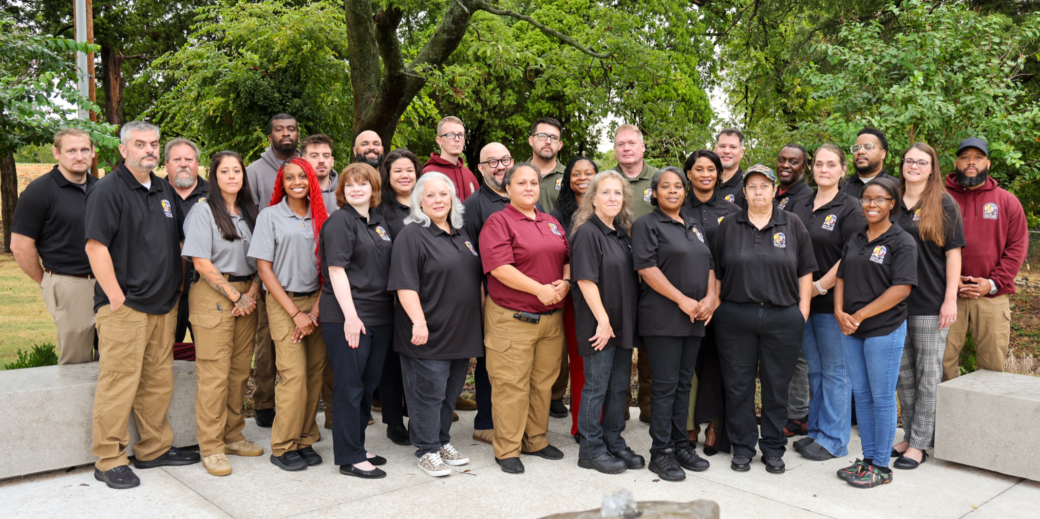 Large group of men and women standing in front of a small water fountain with trees behind them.